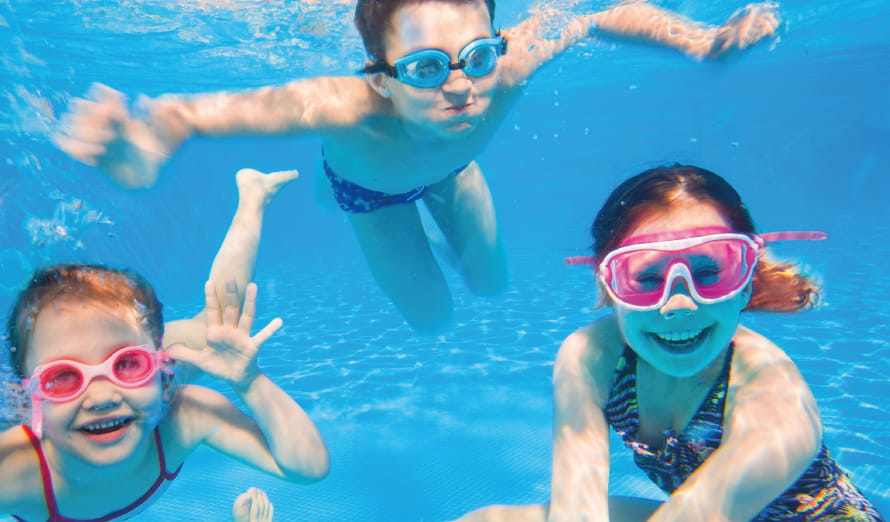 Three young children wearing googles underwater in a blue swimming pool