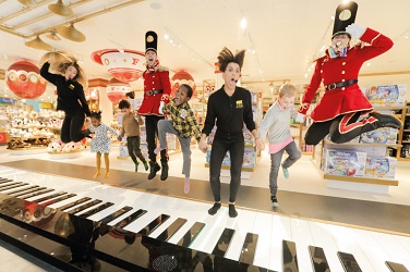 Children, adults and employees dressed as buckingham palace guards jumping on a floor piano at Selfridges UK retail fit out by ISG