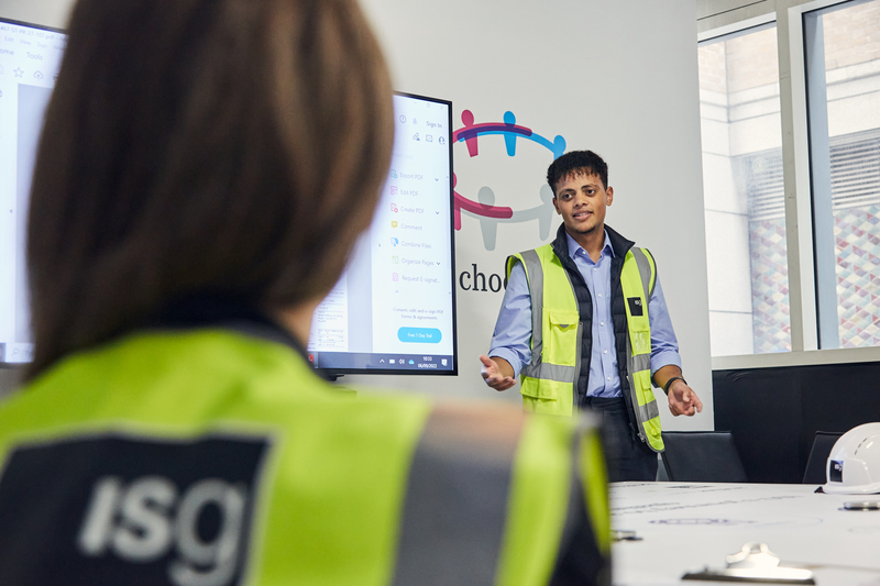 ISG early careers employee in branded high-vis jacket giving a presentation to a colleague in an office